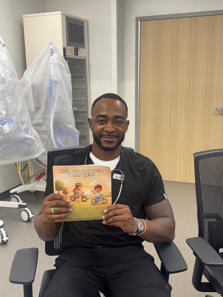 Jovan Balance, RRT seated in a clinical setting holding his book Hey, Big Brother, I Can Help